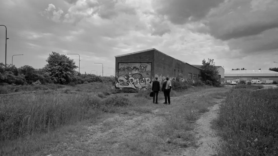 Svartvit scen ur film. Två personer står i ett öde landskap framför en övergiven lokal.
