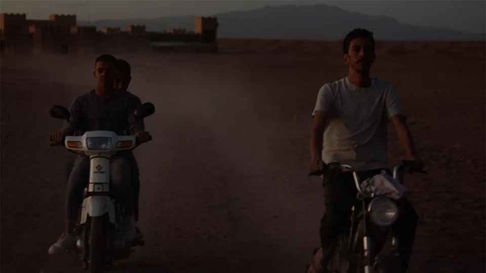Tre unga män åker på två mopeder i ökenlandskap. Scen ur film.