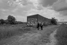 Svartvit scen ur film. Två personer står i ett öde landskap framför en övergiven lokal.