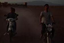 Tre unga män åker på två mopeder i ökenlandskap. Scen ur film.